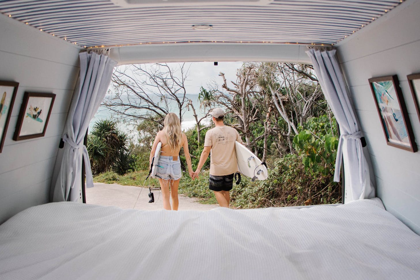 A photo taken from inside a camper van, looking out at a couple walking towards the beach with surfboards.